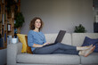 © Rainer Berg/Westend61 - Woman looking away while using laptop sitting on sofa at home