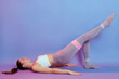© sementsova321 - Photo of woman lying on floor in bridge pose and raising one leg up, using resistance rubber fitness band for training her butt, looking up, wearing leggins and tank top.