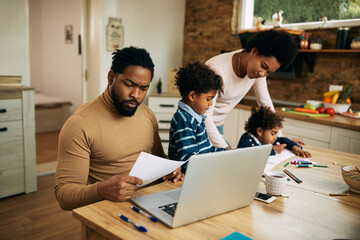 Wall Mural - Black working father analyzing paperwork while mother is assisting their kids with homework at home.