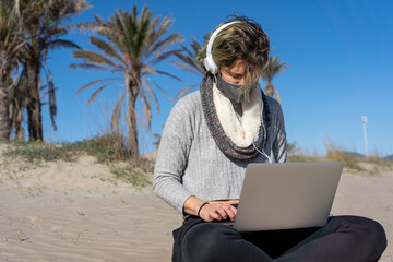 Sticker - Woman wearing a protective mask with headphones working on her laptop on the beach