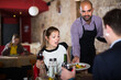 Positive waiter serving meals to young woman with male at cafe