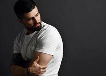 Handsome Modern Brutal Bearded Muscular Man In T-shirt And With Tattoo On Arms Stands Side To Camera Looking Back At Copy Space Over Grey Background. Hipster Stylish Look For Free Lifestyle Concept