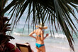 Tropical beach and sea, vacation on sunny paradise coast. Defocused view through palm leaves to woman in blue swimsuit