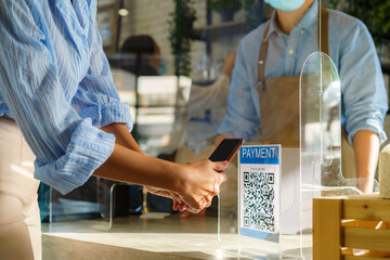 Asian woman customer using mobile scan QR or NFC payment on smart phone paying for her coffee drink in coffee shop and plexi screen and Barista wearing mask in background.