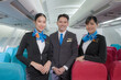 Portrait of three man and woman in blue suit flight attendant/air hostess in economy class cabin smiling to welcome passenger at airplane.