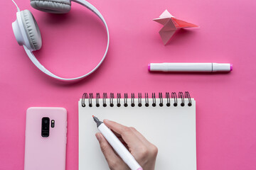 Wall Mural - Hand holding markers with a notebook, headphones, phone, and swan origami on pink background