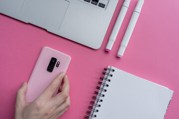 Canvas Print - Top view of markers, notebook, laptop, and hand holding smartphone with pink case on pink background