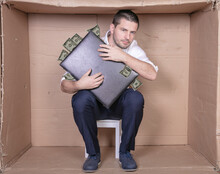 Businessman With A Briefcase Sits In A Tight Cardboard Box Imitating An Office