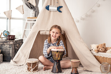 Wall Mural - Little girl playing on traditional african djembe drums sitting in wigwam at home