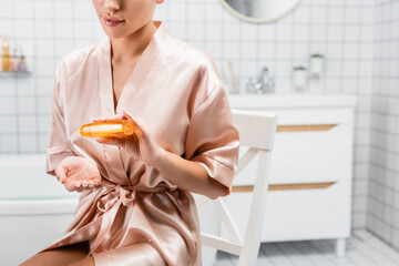 Cropped of woman in silk bathrobe pouring cosmetic oil in bathroom