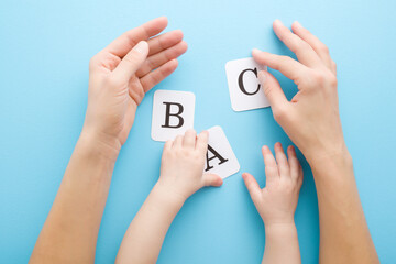 Young adult mother hands showing white cards of letters to baby on light blue table background. Pastel color. Time to learning. Point of view shot. Closeup. Top down view.