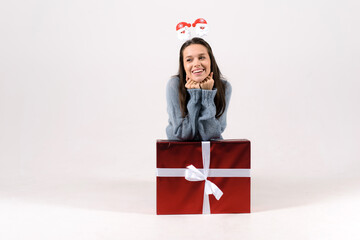 Beautiful excited woman in a blue sweater, sitting near the gift box with white bow, with funy head hoop, isolated on white background