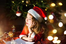 Little Girl In A Santa Hat And Red Dress Under Christmas Tree Is Dreaming, Waiting For The Holiday, Lying On A Plaid Blanket. A Letter On Piece Of Paper, Gifts. New Year, Christmas. Defocus Lights 