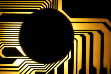 The backlit photo of a yellow and black electrical circuit, with the black connections directed to the center of the circuit, where there is a large round and black component.