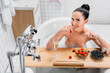  woman smiling while holding strawberry near glass of champagne on wooden tray in bathtub with foam