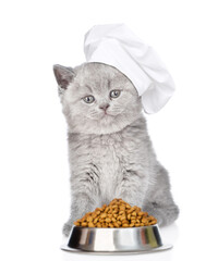 Poster - Kitten wearing chef's hat stands with bowl of dry food. isolated on white background