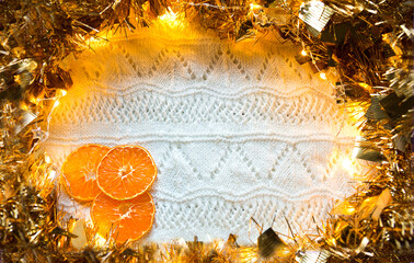 Festive frame made of gold garland with lights and tinsel, slices of dried oranges on a white knitted with a pattern background, copyspace. New year, Christmas, autumn and winter warming atmosphere.