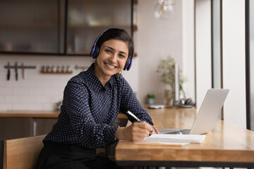 Sticker - Portrait of smiling millennial Indian female student in headphones look at camera studying online on computer. Happy young ethnic woman in earphones make notes work distant on laptop.