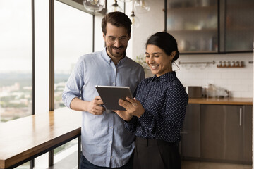 Smiling multiracial young colleagues look at tablet screen discuss financial business online project on gadget. Happy diverse multiethnic coworkers cooperate use pad device at work break in office.