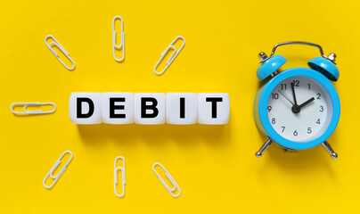 On a yellow background, a blue alarm clock, paper clips and white cubes on which the text is written - DEBIT