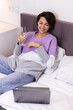 © Анастасия Каргаполов - Happy woman in warm pullover in bed with glass of wine alone by herself resting watching movie comedy smile laugh