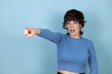 Young caucasian woman over isolated blue background pointing with finger surprised ahead, open mouth amazed expression, something on the front