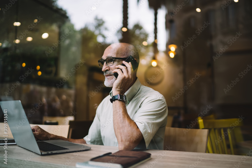 Elderly male freelancer on retirement sitting at coworking desktop with modern netbook technology for doing distance job and making consultancy smartphone talking, aged man in eyewear caling - obrazy, fototapety, plakaty 