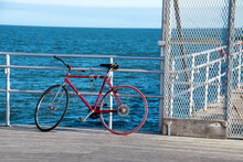 An Old Red Bicycle Leaning Against And Chained To A Fence And Hand Rail On A Wooden Pier With The Blue Ocean In The Background