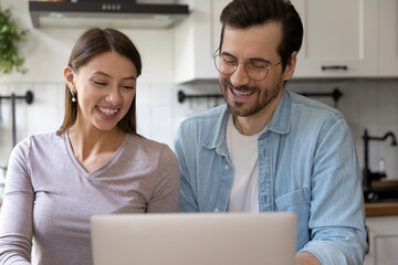 Wall Mural - Close up happy young couple using laptop, sitting in kitchen, smiling wife and husband wearing glasses looking at screen, watching movie or funny video, shopping online, enjoying leisure time