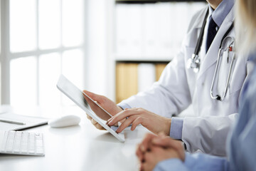 Unknown male doctor and patient woman discussing current health examination while sitting in clinic and using tablet computer, closeup of hands. Medicine and healthcare concept