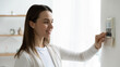© fizkes - Smiling young woman pressing buttons on smart house system close up, standing at home, happy girl customer using air conditioning controller on wall, setting alarm password or comfort temperature