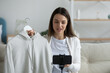 © fizkes - Close up smiling young woman shooting clothing review video, sitting on couch at home, speaking, using smartphone on tripod, girl influencer blogger holding demonstrating clothes to camera