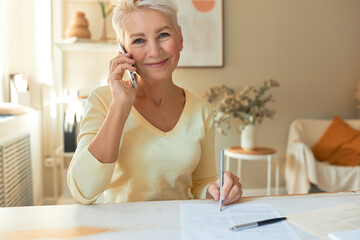 Attractive middle aged female manager sitting at table with documents, looking at camera with pleased smile, holding mobile, making phone call, talking to supplier while working on commercial offer