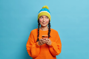 Wall Mural - Thoughtful beautiful teenage girl holds mobile phone and sends text messages thinks what answer to give reads comments under her post in social networks dressed in winter clothes isolated on blue wall