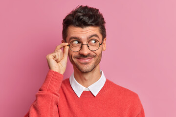 Wall Mural - Headshot of handsome European man with bristle looks aside and smiles pleasantly has thoughtful expression wears round spectacles red jumper isolated over pink background. Human face expressions