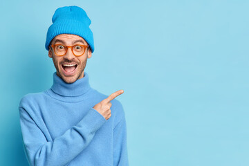 Handsome cheerful Caucasian guy in casual poloneck and hat introduces product demonstrates advertisement points at copy space isolated over blue studio background. You should visit this place