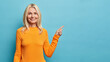 © wayhome.studio  - Waist up shot of blonde middle aged woman with glad expression shows awesome advertisement wears orange jumper isolated over blue background suggests to click link. Your promotional content here
