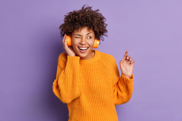 Poster - Joyful teenage girl winks eye and smiles pleasantly has happy mood enjoys listening favorite audio track from playlist wears stereo headphones dressed in orange jumper isolated over purple background