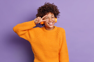 Wall Mural - Positive dark skinned woman makes peace gesture over eye smiles broadly has fun feels amused listens pleasant song in headphones poses against purple background. People and happiness concept