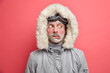 Headshot of emotive puzzled man purses lips and looks aside trembles from low temperature needs to warm wears grey jacket with fur hood poses indoor against pink background. Cold winter time