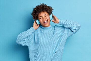 Wall Mural - Carefree joyful dark skinned young woman listens audio track in headphones closes eyes and smiles broadly expresses happy emotions enjoys music isolated over blue background. Monochrome shot