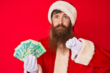 Canvas Print - Handsome young red head man with long beard wearing santa claus costume holding african rand with angry face, negative sign showing dislike with thumbs down, rejection concept