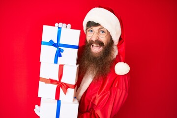 Canvas Print - Handsome young red head man with long beard wearing santa claus costume holding presents smiling and laughing hard out loud because funny crazy joke.