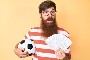 Wall Mural - Handsome young red head man with long beard holding football ball and bunch of dollars clueless and confused expression. doubt concept.