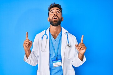 Young hispanic man wearing doctor uniform and stethoscope amazed and surprised looking up and pointing with fingers and raised arms.