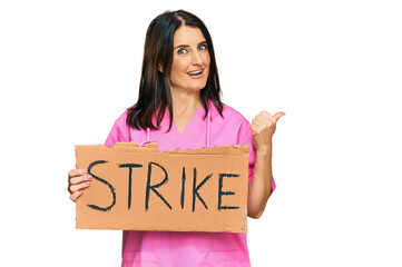 Canvas Print - Middle age brunette doctor woman holding strike banner cardboard pointing thumb up to the side smiling happy with open mouth