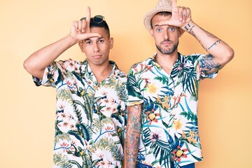 Wall Mural - Young gay couple of two men wearing summer hat and hawaiian shirt making fun of people with fingers on forehead doing loser gesture mocking and insulting.