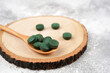 Spirulina tablets in wooden spoon on round stand, light concrete background. Nutrition, vitamin, immunity concept. Close-up, copy space