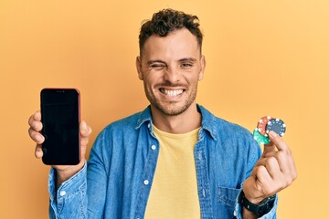 Sticker - Young hispanic man holding poker chips and smartphone showing blank screen winking looking at the camera with sexy expression, cheerful and happy face.