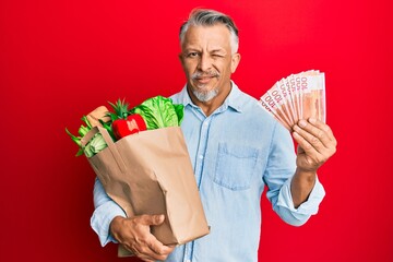 Sticker - Middle age grey-haired man holding groceries and new zealand dollars banknotes winking looking at the camera with sexy expression, cheerful and happy face.